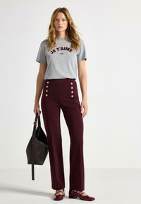 Grå kortærmet t-shirt med "JE T'AIME" i mørkerød, parret med højtaljede burgunderbukser med guldknapper og en sort håndtaske.