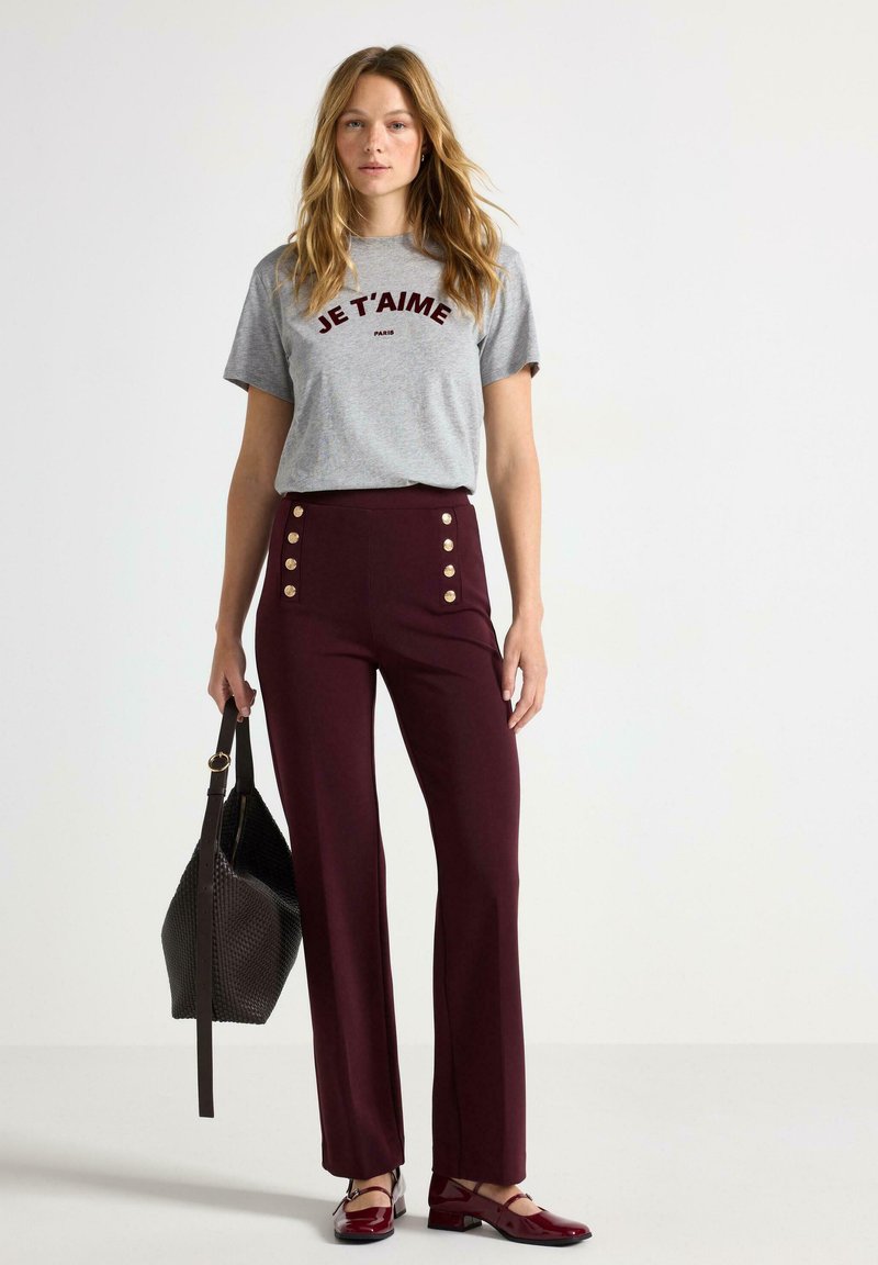 Grå kortærmet t-shirt med "JE T'AIME" i mørkerød, parret med højtaljede burgunderbukser med guldknapper og en sort håndtaske.