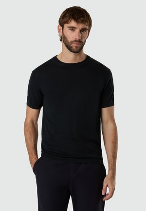 Skæggede mand med brunt hår iført en ensfarvet sort kortærmet skjorte og sorte bukser, stående med den ene hånd i lommen.