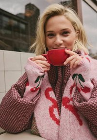 Nertas rožinis megztinis su vyšnių raštais ir raudonomis detalėmis, dėvimas virš žaketo su langeliais rudose tonais. Laiko raudoną keraminį puodelį.