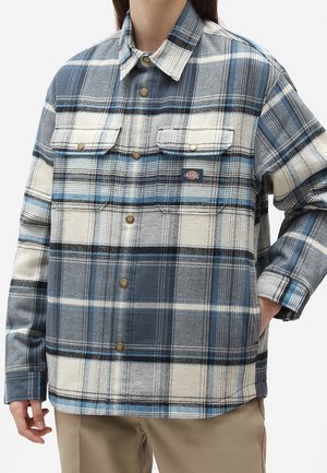 Person wearing blue, white, and gray plaid button-up shirt with two chest pockets and brown buttons, paired with beige pants.