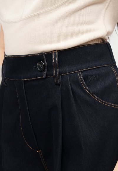 Τζιν με ψηλή μέση από σκούρο denim με αντίθετες ραφές και κλείσιμο με κουμπί, φορεμένα με ανοιχτό μπεζ πλεκτό τοπ.