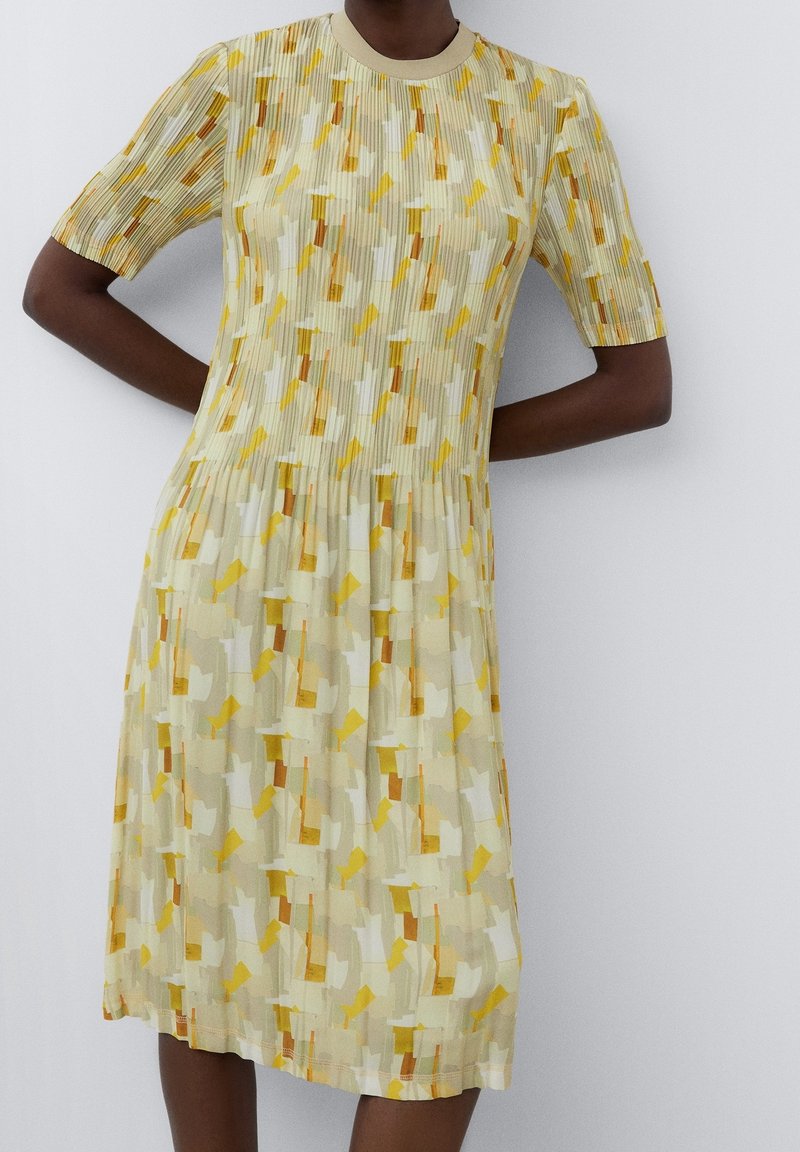 Šaty s krátkým rukávem vyrobené z lehkého, plisovaného materiálu s abstraktním vzorem v žluté, bílé a oranžové barvě a kulatým výstřihem.