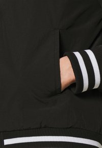 Black bomber jacket featuring ribbed cuffs with white stripes and a side pocket. The material appears smooth with a slight sheen.