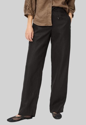 Personne portant un pantalon ample en lin noir, une chemise marron à manches longues boutonnée, et des chaussures noires en maille sans lacets, debout devant un fond uni.