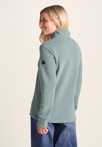 Woman with blonde hair smiling, wearing a light green fleece jacket and blue jeans, facing slightly to the left against a plain background.
