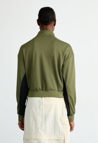 Oliven grønn kort genser med ståkrage, svarte sideakenter og vide ermer, brukt over et lyst beige skjørt.