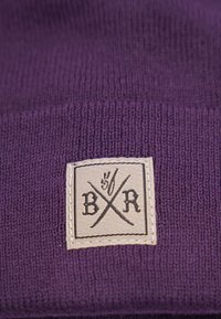 Lila stickad mössa med ett vävt märke med korslagda svärd och texten "B/R". Mjuk textur och rippad design för en snug passform.