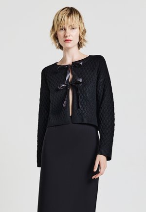Woman wearing a textured black knit cardigan with satin ribbon ties over a long black skirt, standing against a plain light background.