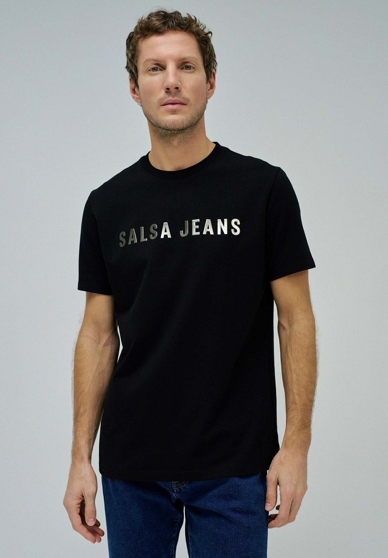 Camiseta de manga corta negra hecha de algodón. Presenta el texto metálico plateado "SALSA JEANS" en el pecho. Cuello redondo clásico.