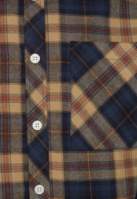 Camisa de franela con un patrón de cuadros en beige, azul marino y óxido; incluye botones blancos y un bolsillo en el pecho izquierdo con diseño a juego.