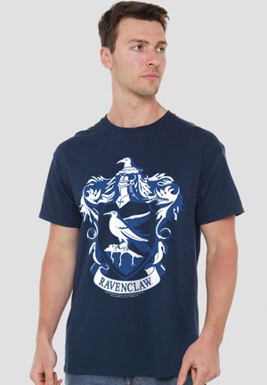 Young man wearing a navy blue T-shirt with the white Ravenclaw crest and text, looking to his right against a light gray background.
