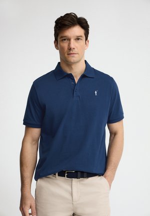 Mężczyzna w granatowej koszulce polo z krótkim rękawem i beżowych spodniach, stojący z jedną ręką w kieszeni na jednolitym tle.