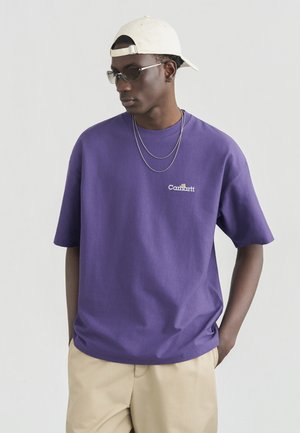 Młody mężczyzna w fioletowej koszulce Carhartt, beżowych spodniach, białej czapce założonej do tyłu, srebrnych łańcuchach i ciemnych okularach przeciwsłonecznych, stojący z rękami w kieszeniach.