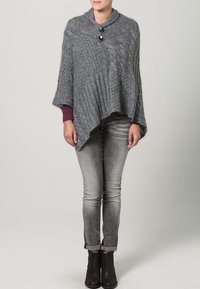 Grå stickad poncho med ribbad textur, med en sjalkrage och två stora svarta knappar. Bärs över en vinröd topp och grå jeans.