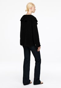 Frau mit blonden Haaren in schwarzer Rüschentbluse, dunklen Schlagjeans und schwarzen Sandalen, die steht und seitlich gegen einen weißen Hintergrund schaut.