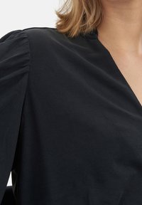 Blouse noire à manches longues avec un décolleté en V et des détails froncés aux épaules, réalisée dans un tissu lisse et léger.