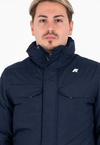 Giacca zip-up blu navy con colletto alto, due tasche frontali e dettagli con logo. Texture liscia e design pratico per calore e stile.