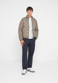 Wattierte Steppjacke in Olivgrün, mit Stehkragen und gerippten Bündchen, kombiniert mit einem grauen Sweatshirt und navyblauen Hose. Weiße Sneakers mit schwarzen Streifen.