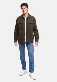Brun, randig button-up jacka med två bröstfickor, bärs över en vit skjorta, tillsammans med blå jeans och vita sneakers.