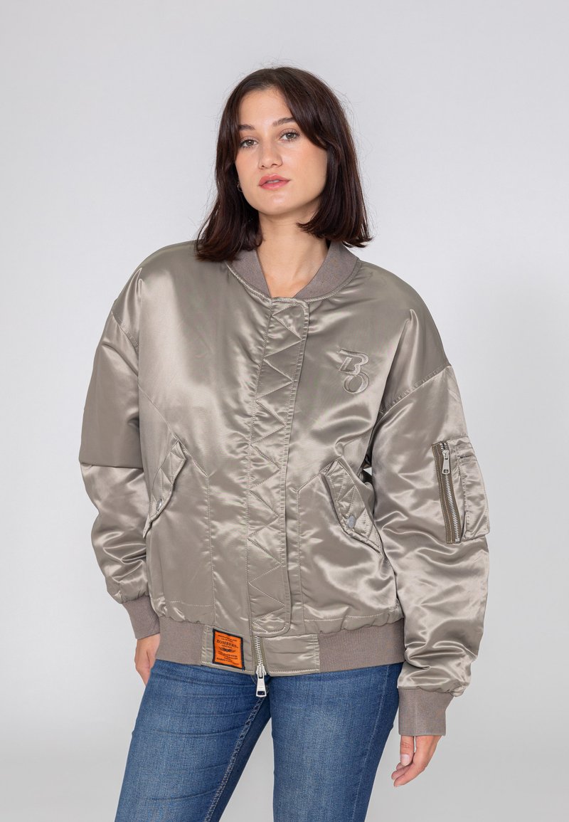 Jasno beżowa satynowa kurtka bomber z prążkowanymi mankietami, zamykanymi kieszeniami po bokach i pomarańczową metką. Posiada wyraźne haftowane logo.