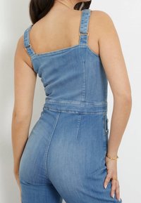 Combinaison en denim avec des bretelles réglables, une coupe ajustée et une couleur bleu clair. Comprend des coutures visibles et des poches latérales.