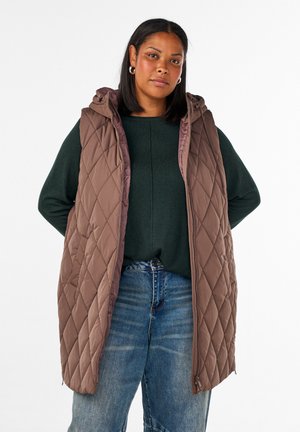 Kvinna med svart mellanlångt hår som bär silverfärgade hoops-örhängen, en mörkgrön tröja, blå jeans och en ärmlös brun quiltad väst med huva.