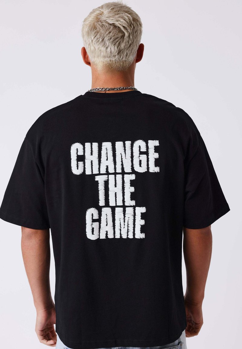 Personne aux cheveux blonds courts portant un T-shirt noir avec le texte blanc en bloc « CHANGE THE GAME » au dos, debout devant un fond uni.