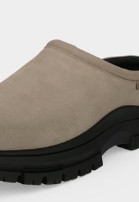 Μπεζ σουέντ παπούτσι slip-on με χοντρή μαύρη λαστιχένια σόλα και βαθύ ανάγλυφο πάτο, γωνιακά τοποθετημένο για να δείχνει πλαϊνές και μπροστινές λεπτομέρειες.