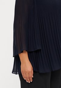 Marineblå plisserte topp med gjennomskinnelig stoff, med luftige ermer og et flytende design. Nærbilde av hånd som hviler på det tettsittende bunnen.
