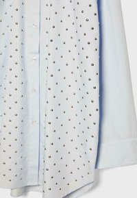 Svetlomodrá košeľa s dlhým rukávom, predným zapínaním na gombíky a striebornými kovovými nitmi rovnomerne pokrývajúcimi jednu stranu predného panelu.