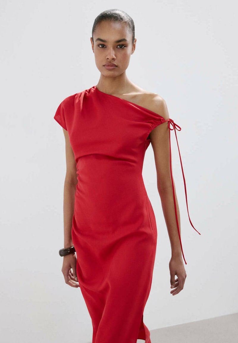 Femme portant une robe rouge ajustée à bretelle unique avec une lanière nouée sur l'épaule découverte, debout devant un fond clair uni.
