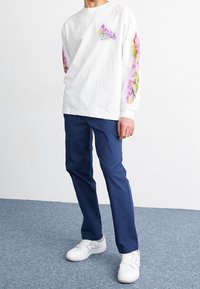 Camicia bianca a maniche lunghe con design astratto colorato sulle maniche, abbinata a pantaloni blu navy e sneakers bianche su un pavimento in moquette blu.