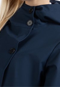 Chaqueta de lluvia azul marino con un alto cuello, botones negros brillantes y una tela suave y lisa con detalles de costura mínimos.