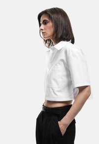 Witte geknipte shirt met korte mouwen, knoopsluiting vooraan en borstzak. Draagt bij hoge taille zwarte broeken en een zwarte riem.