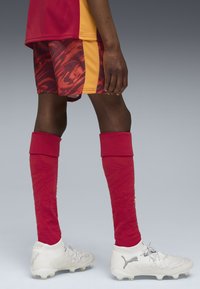 Maglietta rossa e gialla con tessuto testurizzato, abbinata a pantaloni corti rossi a fantasia, calzini rossi al ginocchio e scarpe bianche con logo PUMA.