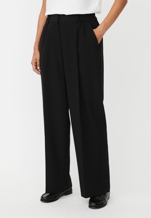 Vrouw met hoge taille zwarte wijde broek met zijzakken en zwarte loafers, gecombineerd met een wit, ingestopt shirt.