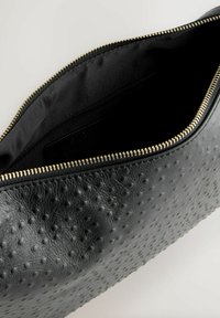 Musta nahkakotelo, jossa on teksturoitu pinta ja vetoketjusuljin. Sisäpuoli on vuorattu mustalla kankaalla, jossa on lisävetoketjutasku. Kultaiset vetoketjun yksityiskohdat.