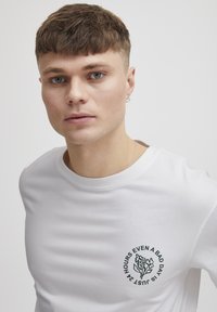 Hvid bomulds t-shirt med korte ærmer, featuring et rundt grafik på venstre bryst med sort tekst og blomsterillustration.