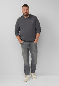 Uomo con barba e capelli raccolti indossa una felpa grigia scura, jeans grigi sbiaditi e sneakers bianche, in piedi con le mani in tasca contro un muro grigio.