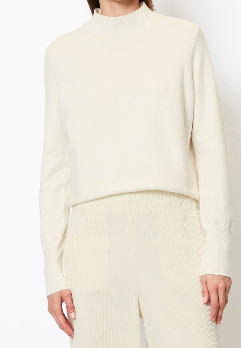Femme portant un pull à col roulé crème à manches longues, rentré dans un pantalon côtelé crème taille haute, debout devant un fond blanc uni.