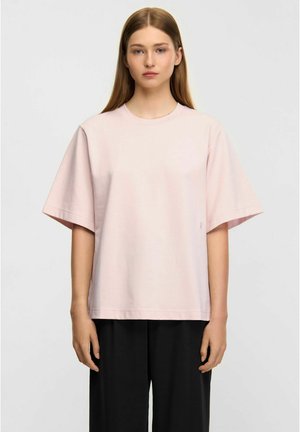 Ung kvinde med glat hår iført en løs, lyserød kortærmet skjorte og løse sorte bukser, stående mod en lys baggrund.