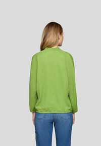 Frau, die von hinten zu sehen ist, trägt ein hellgrünes, strukturiertes Langarmoberteil und blaue Jeans mit Verzierungen auf dem linken Oberschenkel, vor einem schlichten grauen Hintergrund.