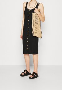 Czarna sukienka z prążkowanego dżerseju z guzikami z przodu, w zestawieniu z plecioną beżową torbą i czarnymi sandałami. Regulowane ramiączka i długość midi.
