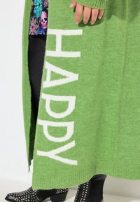 Cárdigan verde de punto con la palabra "HAPPY" en letras blancas; presenta puños y dobladillo acanalados, combinado con botas de tobillo negras con tachuelas.