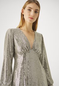Robe en sequins argentés avec des manches longues, décolleté en V et surface texturée. Elle présente un corsage ajusté et une jupe fluide.