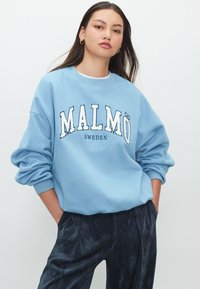 Világoskék pulóver "Malmö" és "Svédország" kontrasztos fehér felirattal. Laza szabású, leejtett vállrésszel és bordázott mandzsettákkal.