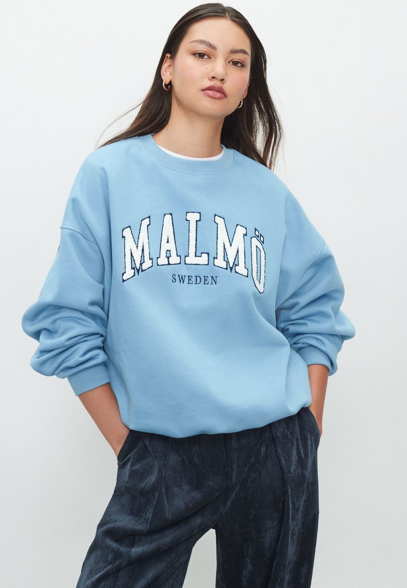 Világoskék pulóver "Malmö" és "Svédország" kontrasztos fehér felirattal. Laza szabású, leejtett vállrésszel és bordázott mandzsettákkal.