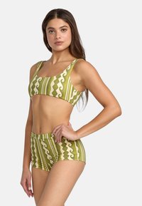 Green and beige two-piece swimsuit featuring geometric patterns and stripes. The top has wide straps; the bottoms are shorts with a fitted design.