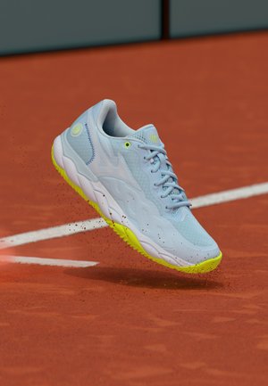 Pantof sport albastru deschis cu talpă galben neon aruncând lut roșu pe un teren de tenis lângă linii albe de delimitare.